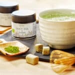 Matcha thee en poeder, biologische ceremoniële matcha in potten, met houten lepel en kookschaal op een zwart leisteen ondergrond.