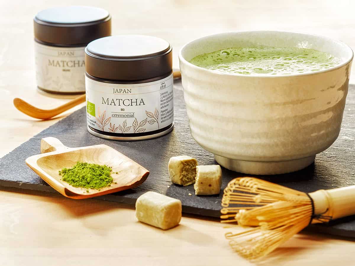 Matcha thee en poeder, biologische ceremoniële matcha in potten, met houten lepel en kookschaal op een zwart leisteen ondergrond.