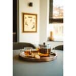 Herbruikbare theepot met theelicht en koekje op houten dienblad, uitnodigend interieur met natuurlijk licht, ontspannen sfeer, perfect voor thee en koffie bij De Heeren van 17.