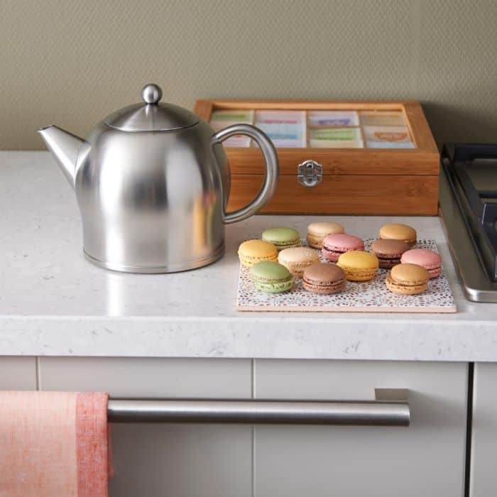Delicate pastelgekleurde Macarons op een witte doily naast een stalen theepot op een keukenblad met taartdoos en oven – perfecte chocolade- en fruitige snoepjes voor een verfijnd dessert.