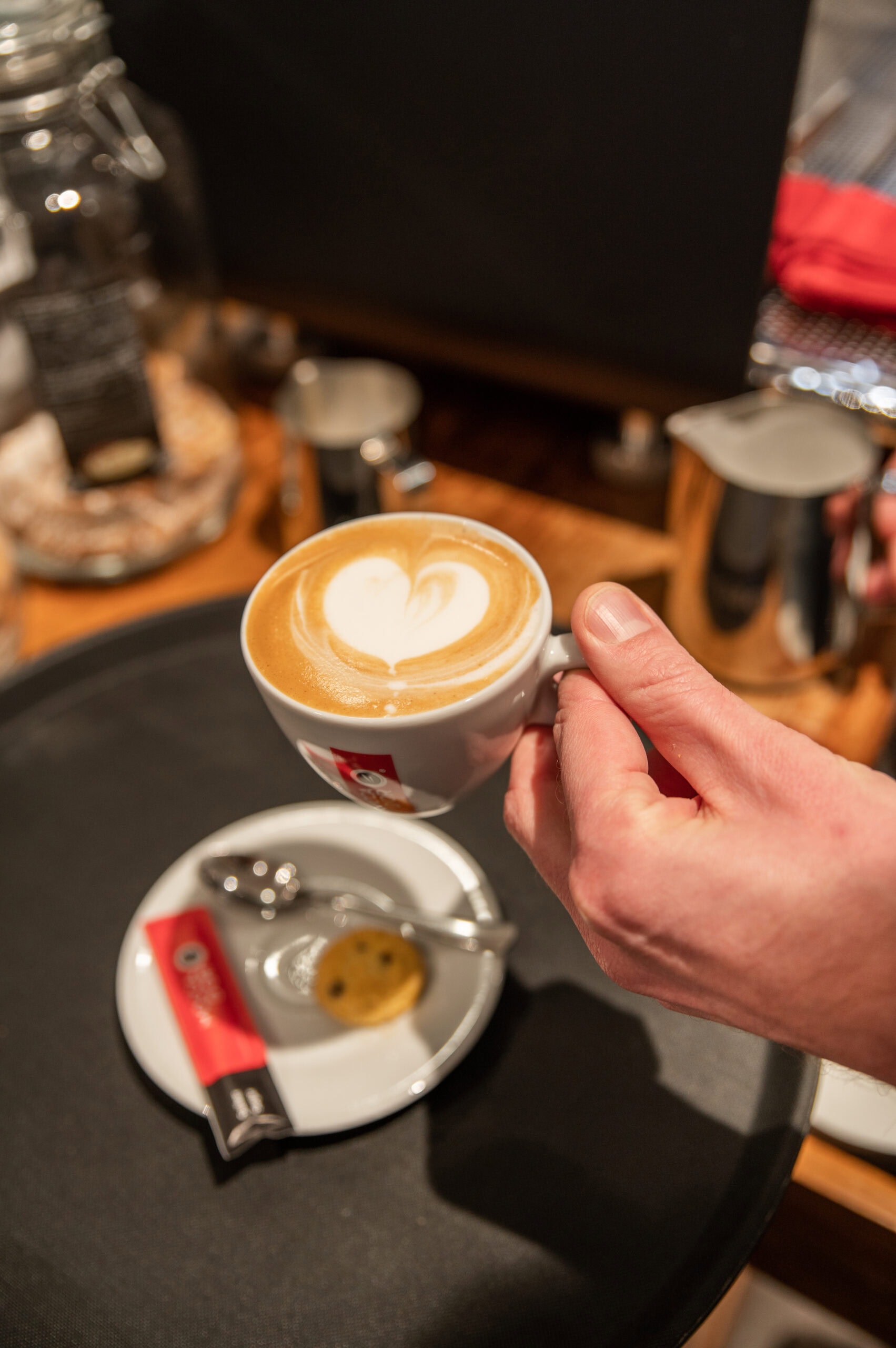 Sfeervolle foto van een hand die een kopje cappuccino met hartvormige latte art vasthoudt bij De Heeren van 17. Gezellig koffieadres in Nederland met ambachtelijke koffie en huisgemaakte lekkernijen.