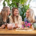 Gezellige vriendinnen genieten van high tea met tea pots en macarons aan een houten tafel, sfeerfoto met groene planten en natuurlijk licht, perfect voor De Heeren van 17.