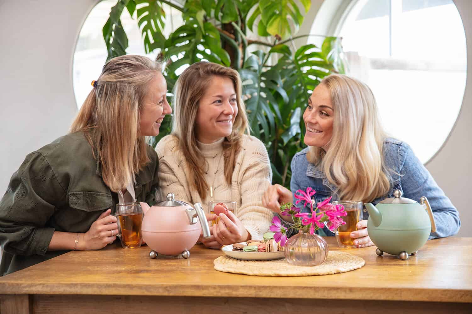 Gezellige vriendinnen genieten van high tea met tea pots en macarons aan een houten tafel, sfeerfoto met groene planten en natuurlijk licht, perfect voor De Heeren van 17.
